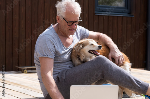 An elderly man is playing with a dog sitting on the terrace and working at a computer