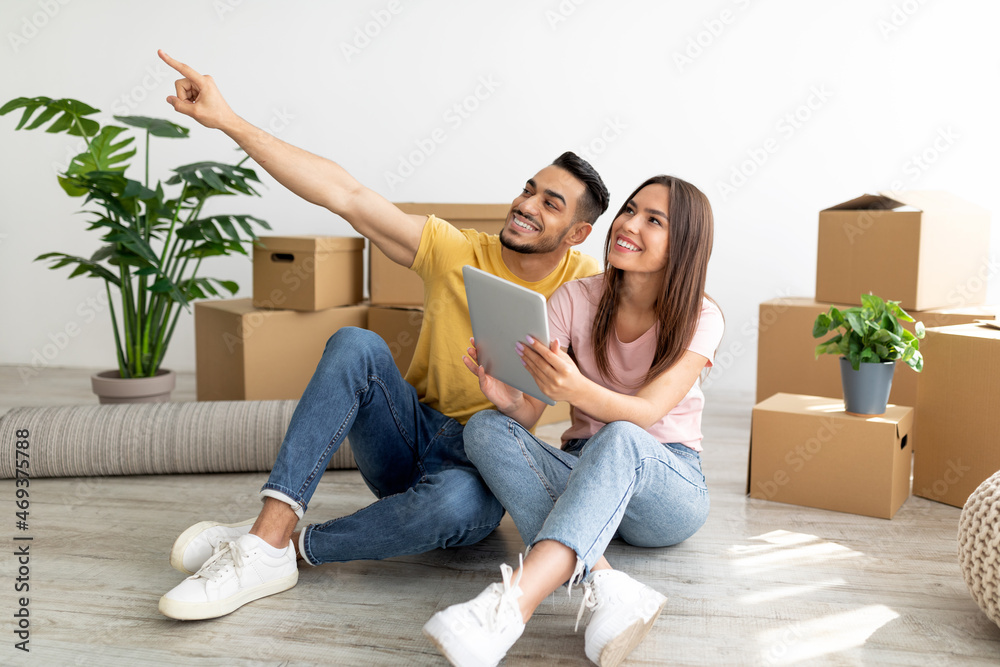 Happy young interracial couple with tablet sitting on floor among boxes, planning furniture placement in new apartment Happy young interracial couple with tablet sitting on floor among boxes, planning furniture placement in new apartment