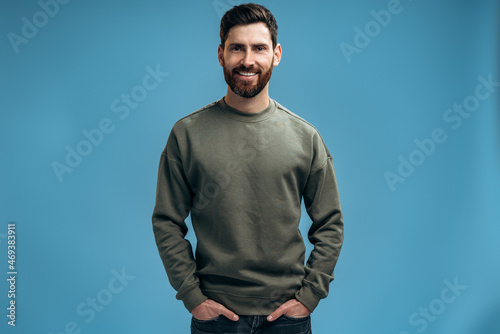 Portrait of serious bearded young man standing and looking at the camera with calm confident attentive expression, worker or employee in casual outfit. Studio shot isolated on blue background