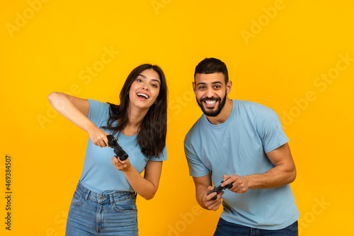Having fun. Playful arab couple playing video games together, holding and using joysticks over yellow background