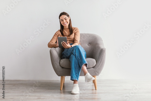 Positive young woman working online, sitting in armchair with digital tablet against white studio wall, full length