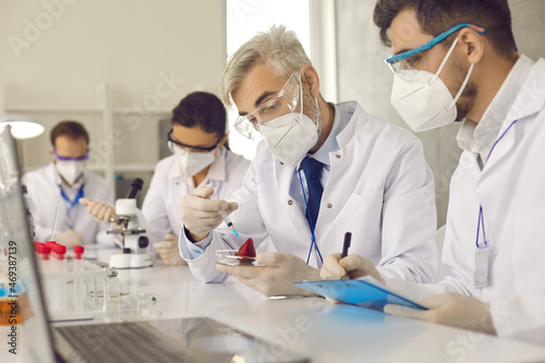 Team of research scientists in protective respiratory face mask and sterile white coat working lab equipment and computer inspecting strawberry on nitrates in laboratory. Food quality control