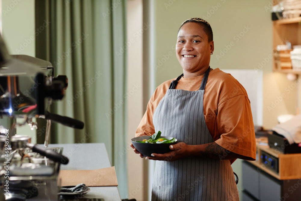 Horizontal medium portrait of cheerful young African American woman with stylish short hair wearing apron standing in cafe kitchen holding bowl with salad smiling at camera.. Horizontal medium portrait of cheerful young African American woman with stylish short hair wearing apron standing in cafe kitchen holding bowl with salad smiling at camera..