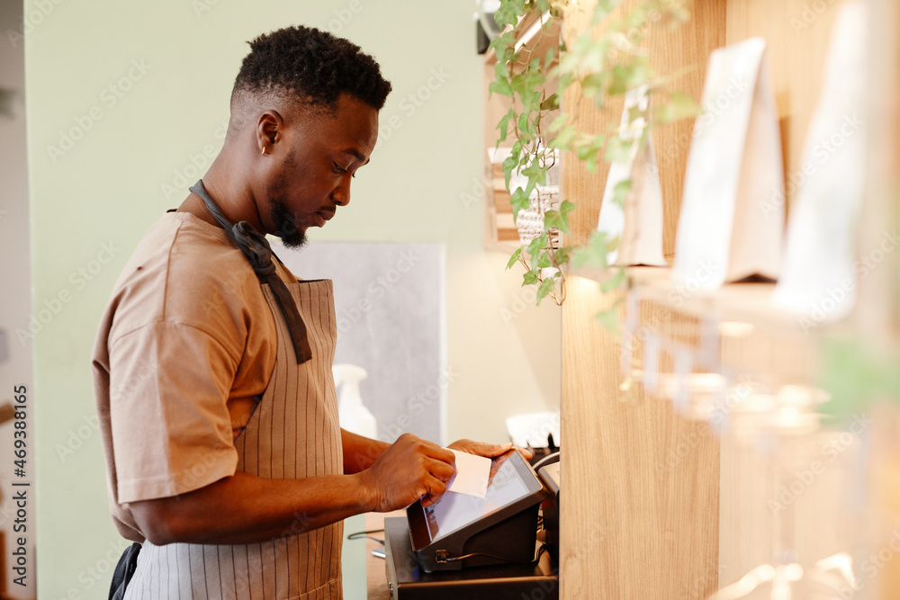 Fototapeta premium Horizontal side view shot of modern small cafe worker working with customer taking order using cash register