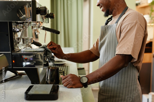 Horizontal side view shot of young African American man working as barista in small cafe making espresso using coffee machine