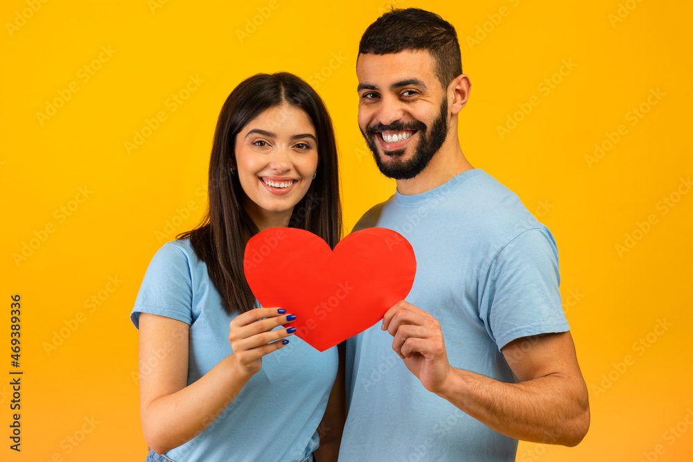 Valentine's day. Loving arab spouses embracing and holding heart, smiling to camera over yellow studio background