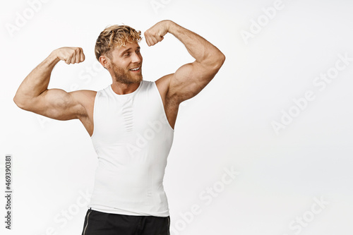 Handsome athlete looking strong, flexing biceps, showing his strong muscles arms, smiling pleased, workout and doing fitness exercises, white background