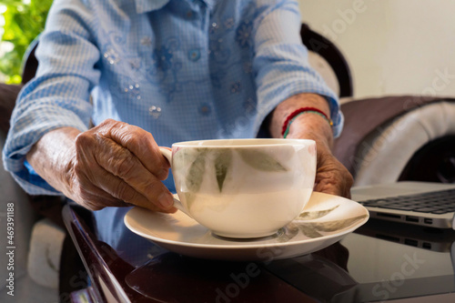 Older woman's hands drinking cup of tea with laptop working.