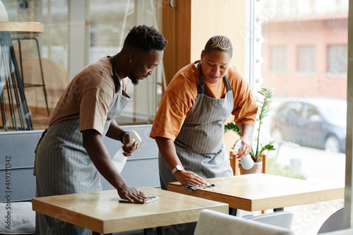 Young African American man and woman wearing aprons working in small cafe preparing tables for customers cleaning them with spray detergent and chatting