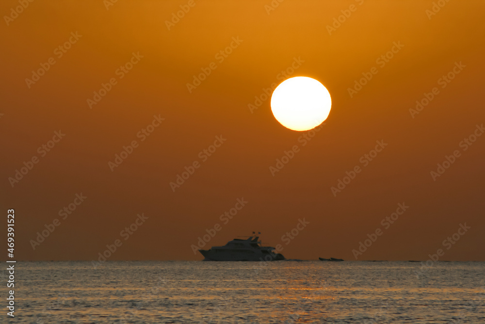 Seascape with silhouette of a pleasure ship and disk of the sun in the red sky.