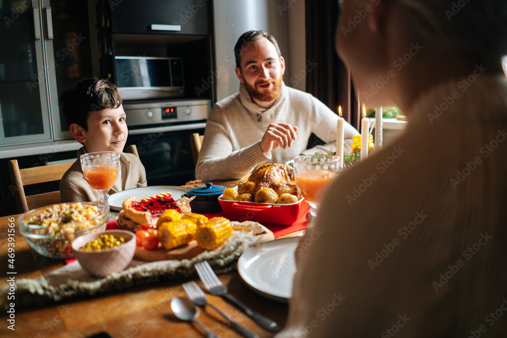 View from back of happy young family enjoying at festive Christmas table during holiday family party. Cheerful mother, father and little son having dinner xmas party, eating and chatting at home.