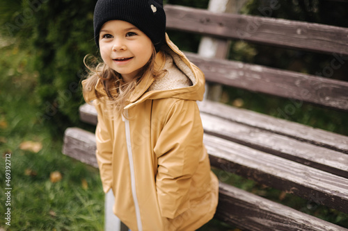 Happy cute little girl in jacket and hat posing to photographer outdoor by the bench. Little model have fun in autumn time