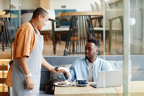 Young Black man having lunch in modern cafe sitting at table paying for order with bank card photo