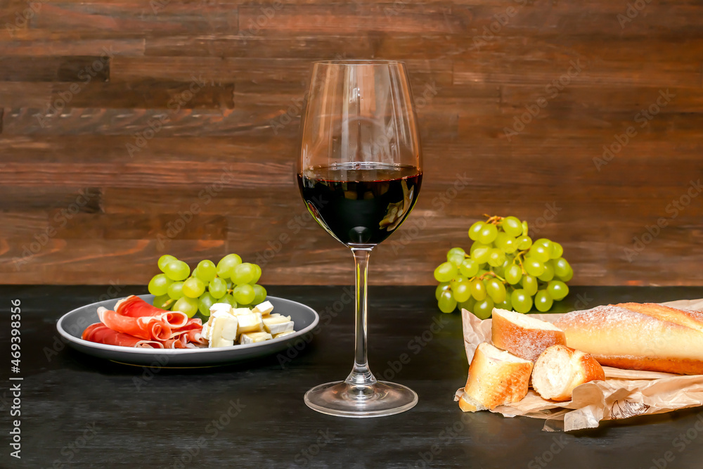 Red wine in glass, brie cheese, blue cheese, baguette, bread, grapes, jamon meat, salami on plate on black wooden table, alcohol drink with snacks, still life close up Red wine in glass, brie cheese, blue cheese, baguette, bread, grapes, jamon meat, salami on plate on black wooden table, alcohol drink with snacks, still life close up