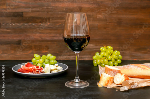 Red wine in glass  brie cheese  blue cheese  baguette  bread  grapes  jamon meat  salami on plate on black wooden table  alcohol drink with snacks  still life close up