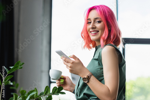 cheerful businesswoman with pink hair and piercing holding cup of coffee and smartphone. photo
