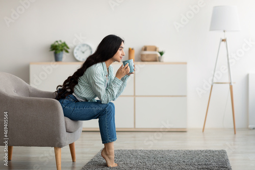 Tableau sur toile Side view of young Indian woman sitting in armchair, sipping hot coffee, enjoyin
