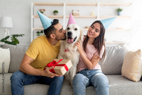 Young multinational couple in party hats celebrating dog's birthday, holding gift box, kissing their pet at home