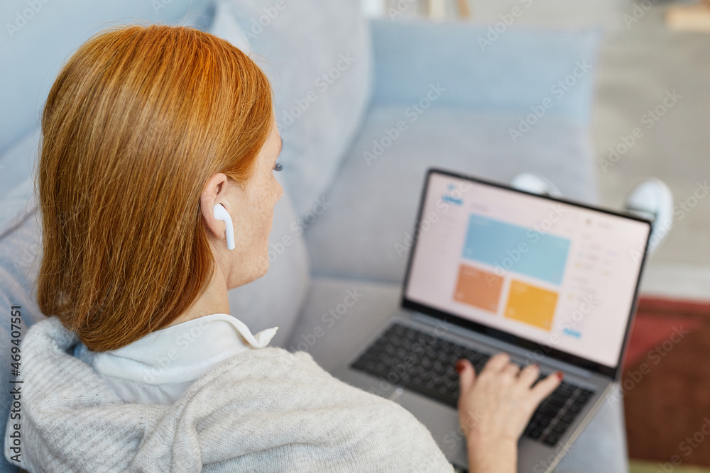 Back view portrait of adult red haired woman using laptop on sofa while studying online from home, copy space Back view portrait of adult red haired woman using laptop on sofa while studying online from home, copy space