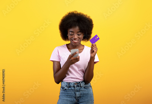 Money transfer and online payment. Happy black lady holding debit credit card and using smartphone, yellow background