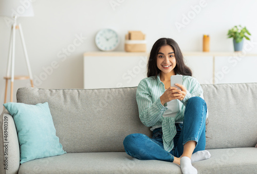 Charming young Indian woman sitting on sofa with smartphone, checking social media, chatting online at home
