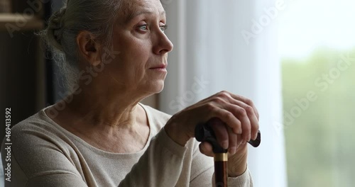 Cropped close up of depressed hoary older adult female living with physical disability sit by window alone hold stick. Retired old woman with walking difficulties look at distance feel weak hopeless