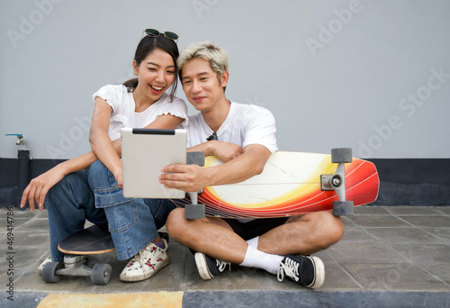 Young asian couple sit resting after skateboarding, take a selfie. Young man in a white T-shirt holding tablet computer while the woman push finger on the screen. photo