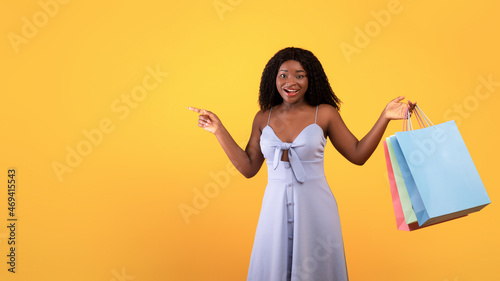 Cheerful young black woman with colorful shopping bags pointing aside, offering big sale or promo on orange background