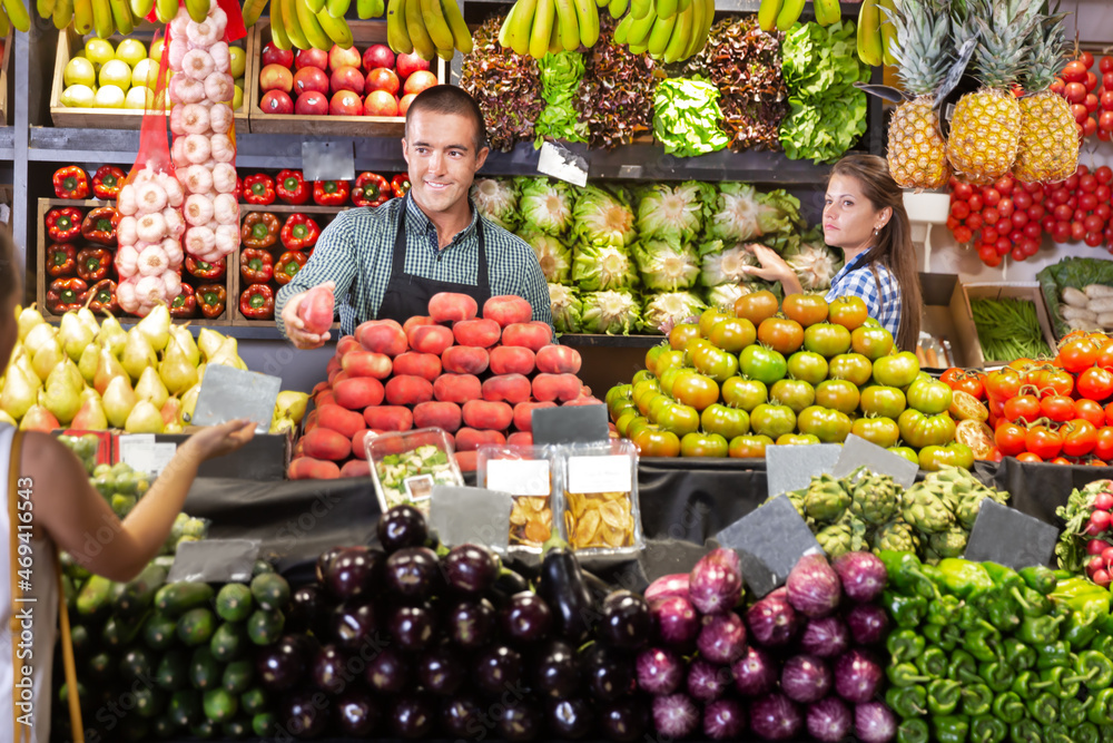 Two friendly shop assistants behind counter selling fruits and vegetables to female buyer
