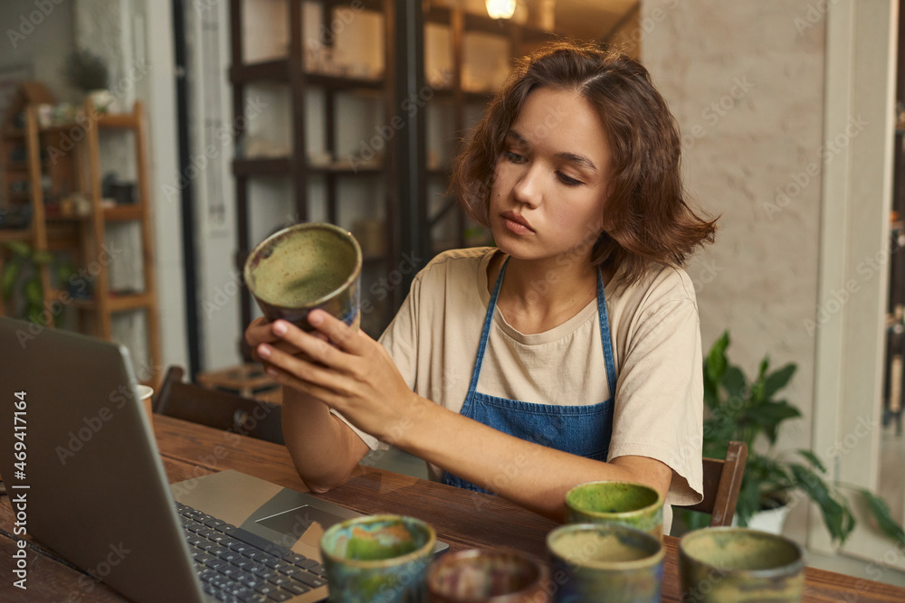 Young creative craftswoman in workwear holding selfmade clay mug and looking at it by her workplace Young creative craftswoman in workwear holding selfmade clay mug and looking at it by her workplace