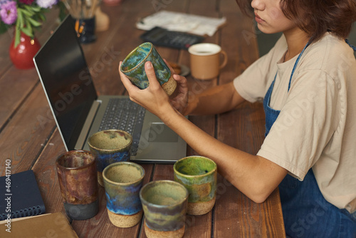 Young pitcher sitting by table in front of laptop while holding earthenware and thinking of ideas about painting handmade mugs