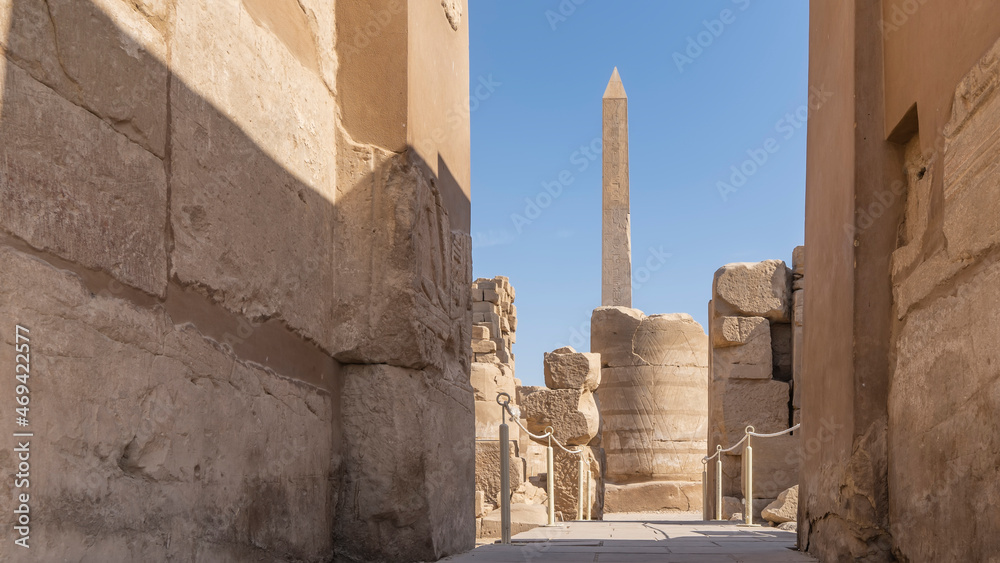 The ancient Karnak Temple in Luxor. In the passage between the stone walls, a dilapidated column and a tall obelisk are visible against the blue sky. Egypt