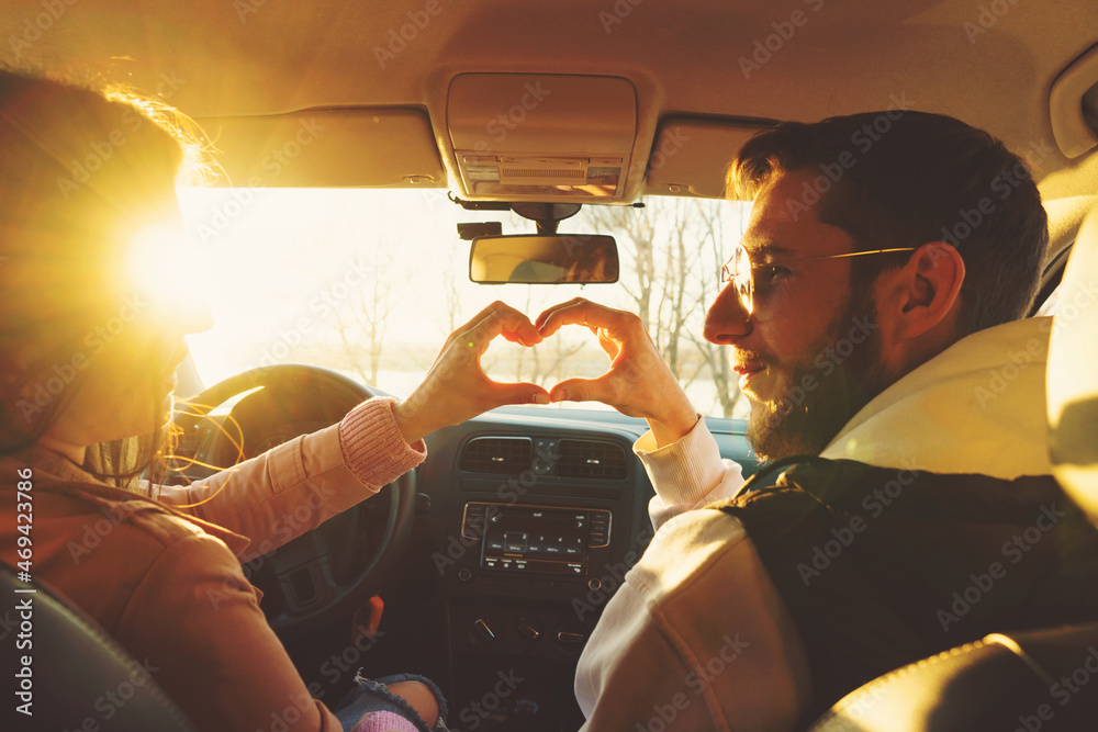 Romantic couple makes heart shape with their hands in car. Female and male hands make up heart shape. Riding sunny day. View from rear seat. Road trip concept. Woman driving - obrazy, fototapety, plakaty 