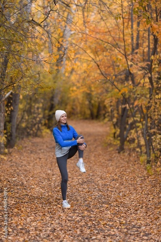 Outdoor pursuit. Caucasian fit woman in blue sportswear and a knit cap warning up her knees before jogging in the forest in autumn, vertical photo, selective focus © junky_jess