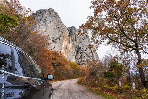 Travel in the mountains by car. Black SUV rides on a road among yellow autumn deciduous trees next to huge mountains of white stone © Sergey + Marina