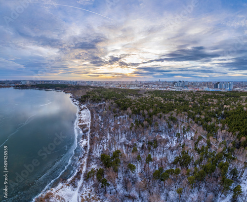 Snow-covered forest on lake shore with the first ice at sunset and the city on horizon, auerial view