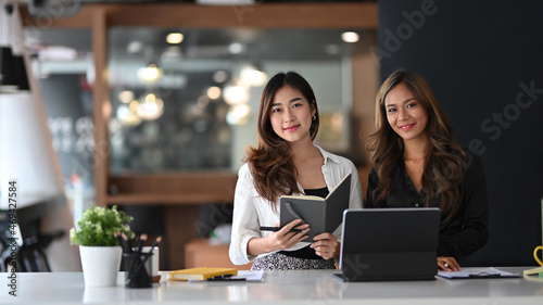 Two beautiful businesswoman sitting together in modern office and smiling to camera.