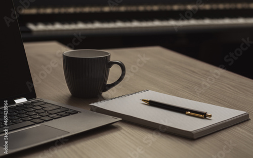 Close-up pencil on a notebook next to a laptop and cup of coffee on a beige wood table against the background of a piano. Horizontal view. Pencil tip in focus.
