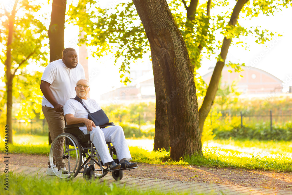 Caregiver and old man in a wheelchair. Professional nurse and patient walking outdoor in the park at sunset. Assistance, rehabilitation and health care. Caregiver and old man in a wheelchair. Professional nurse and patient walking outdoor in the park at sunset. Assistance, rehabilitation and health care.