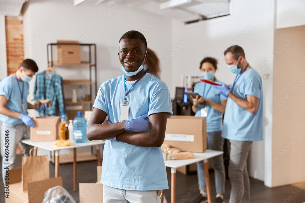 Fototapeta premium Portrait of smiling african american young male volunteer in blue uniform, protective mask and gloves standing with arms crossed. Team sorting, packing food stuff in cardboard boxes
