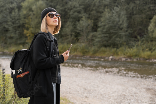 Caucasian girl tourist traveling near the river with a backpack, nature background, stylish young woman traveler in a black coat, side view.
