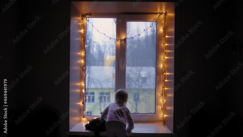 Child Looking out Window During Snow. A little baby at Christmas night. Girl looking through the window. A child stands at the window against the background of Christmas decorations. A lonely child.