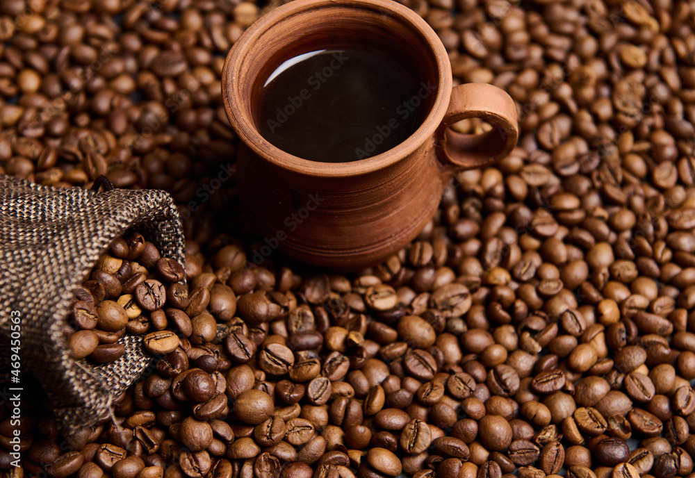 Fototapeta premium High angle view of a clay coffee cup with freshly brewed coffee drink with steam on the background of a bag with grains and freshly roasted coffee beans . Copy space for advertising. Still life