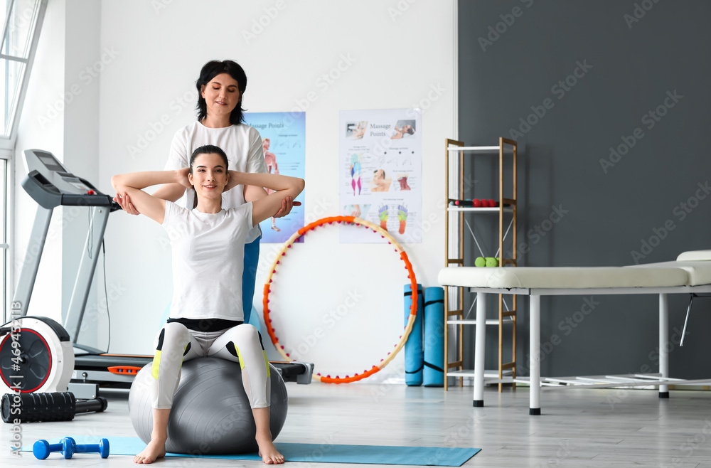 Fototapeta premium Mature physiotherapist working with young woman on fitball in rehabilitation center