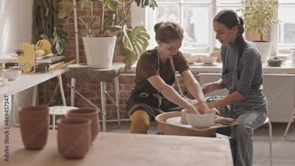 Slowmo shot of two young women sitting next to pottery wheel in cozy workshop, working with clay and having conversation Slowmo shot of two young women sitting next to pottery wheel in cozy workshop, working with clay and having conversation