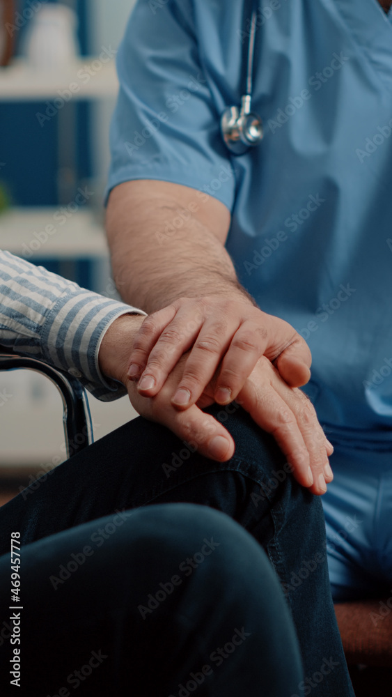 Close up of hands of man nurse comforting senior patient with chronic disability in nursing home. Medical assistant giving support to person sitting in wheelchair. Specialist helping adult