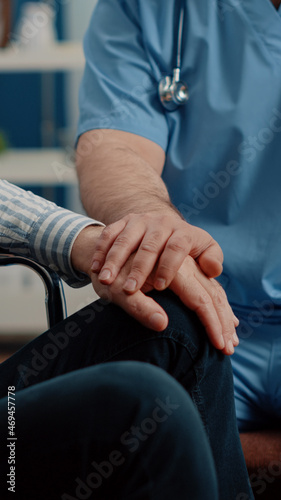 Close up of hands of man nurse comforting senior patient with chronic disability in nursing home. Medical assistant giving support to person sitting in wheelchair. Specialist helping adult