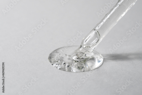 Transparent facial serum in glass pipette pouring on grey background. Drop of liquid gel with hyaluronic acid closeup. Cosmetic product macro. Copy space.