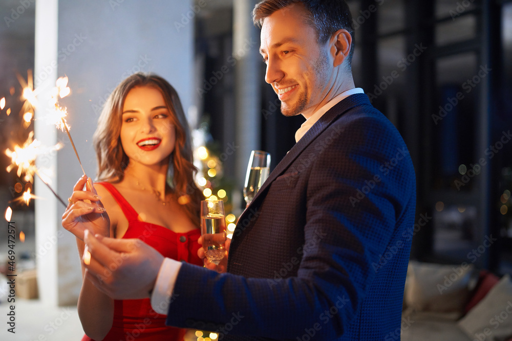 Handsome man in suit and beautiful woman in red dress holding sparklers and glass of champagne. Young couple enjoying christmas evening together at home. Handsome man in suit and beautiful woman in red dress holding sparklers and glass of champagne. Young couple enjoying christmas evening together at home.