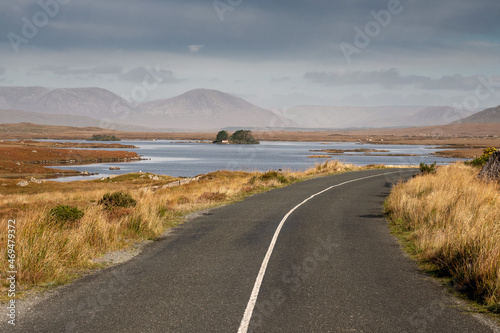 Small narrow asphalt road view view on stunning scenery in the background with lake with small island and big tall mountains. Connemara, Ireland. Irish landscape scene. Travel and tourism concept